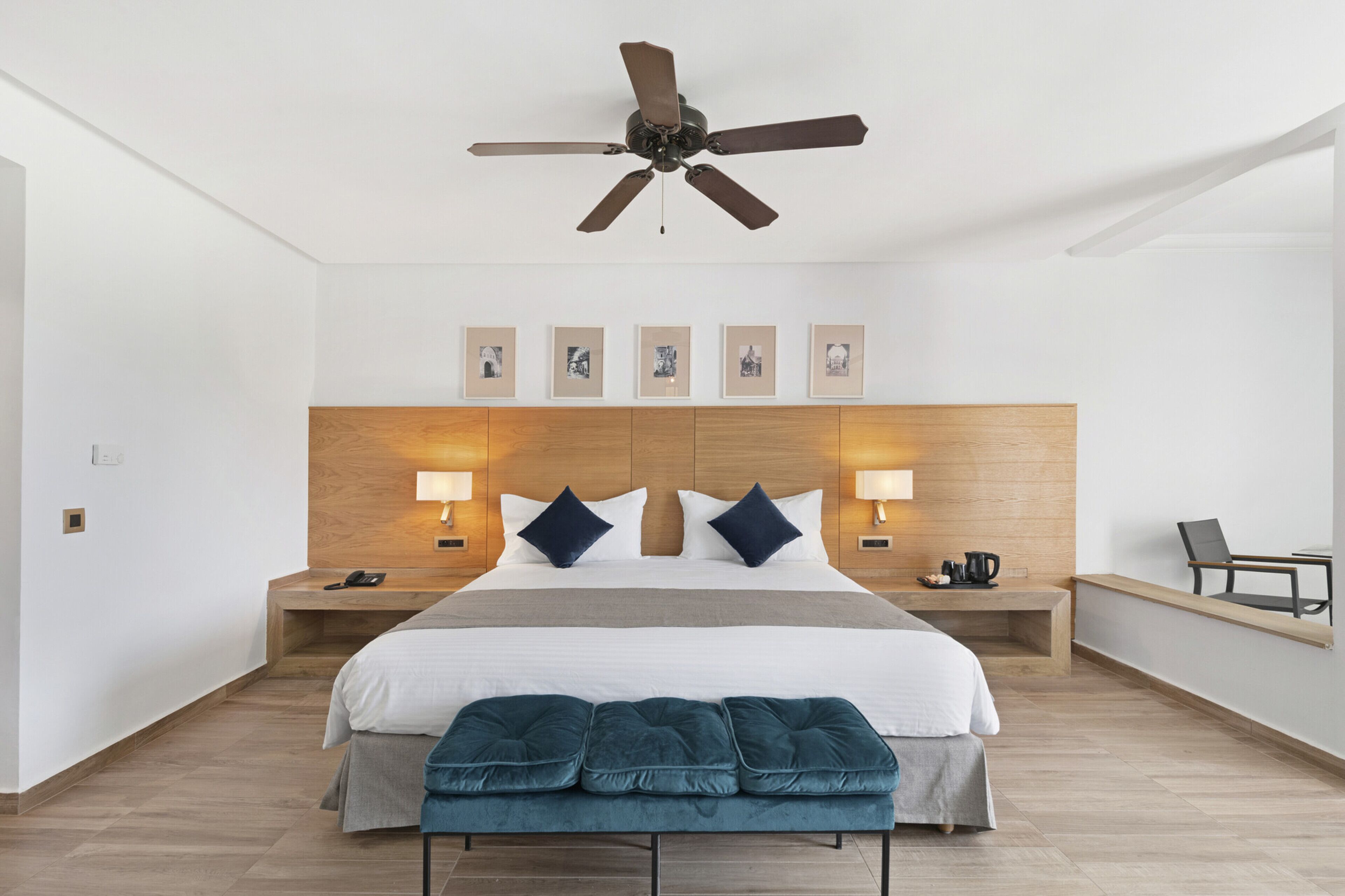 Una habitación de hotel moderna con una cama grande, un ventilador de techo, lámparas de noche, un cabecero de madera, un banco y cuadros en la pared.