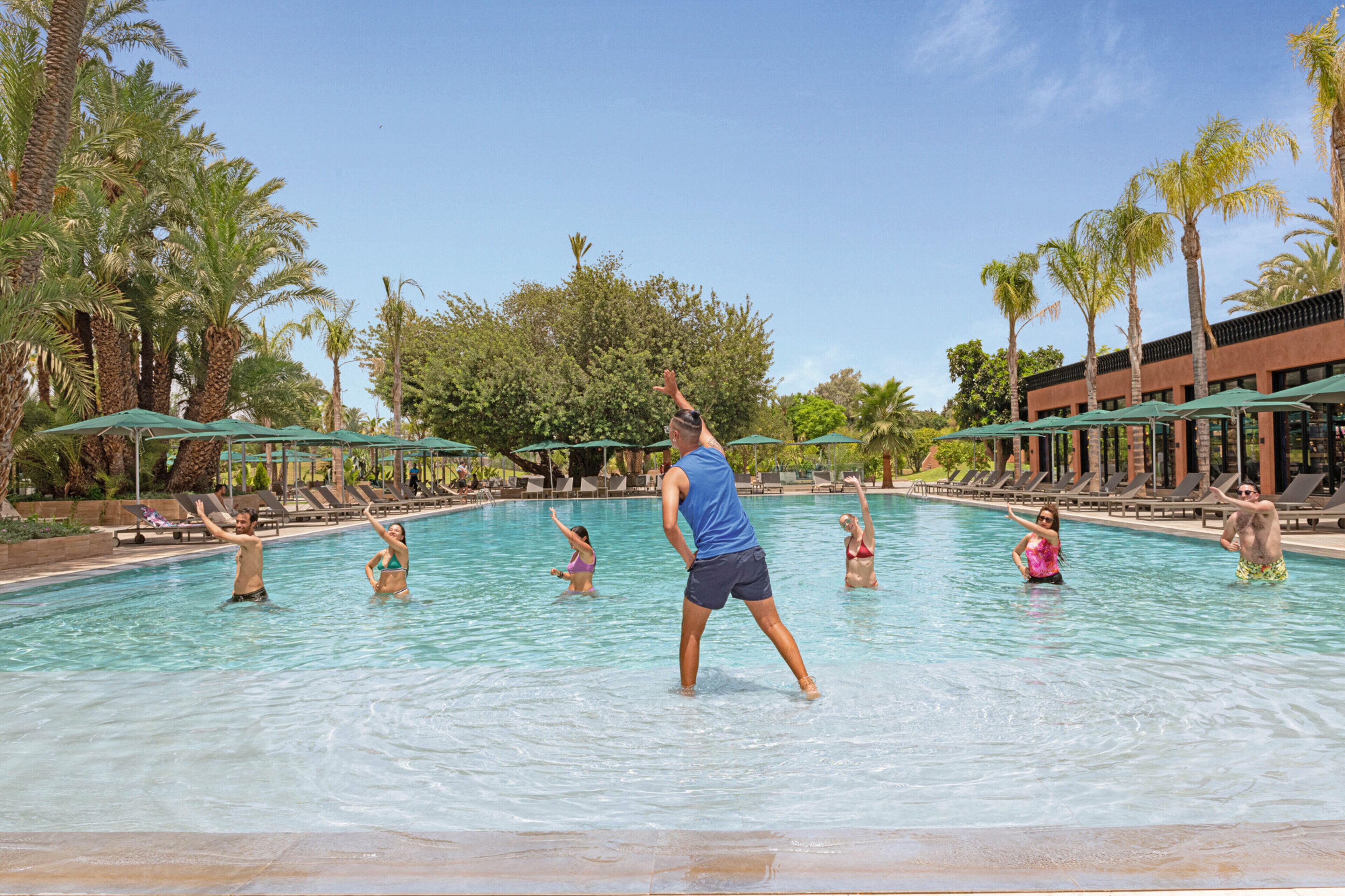 Una piscina con gente disfrutando del agua, rodeada de palmeras y sillones de descanso.