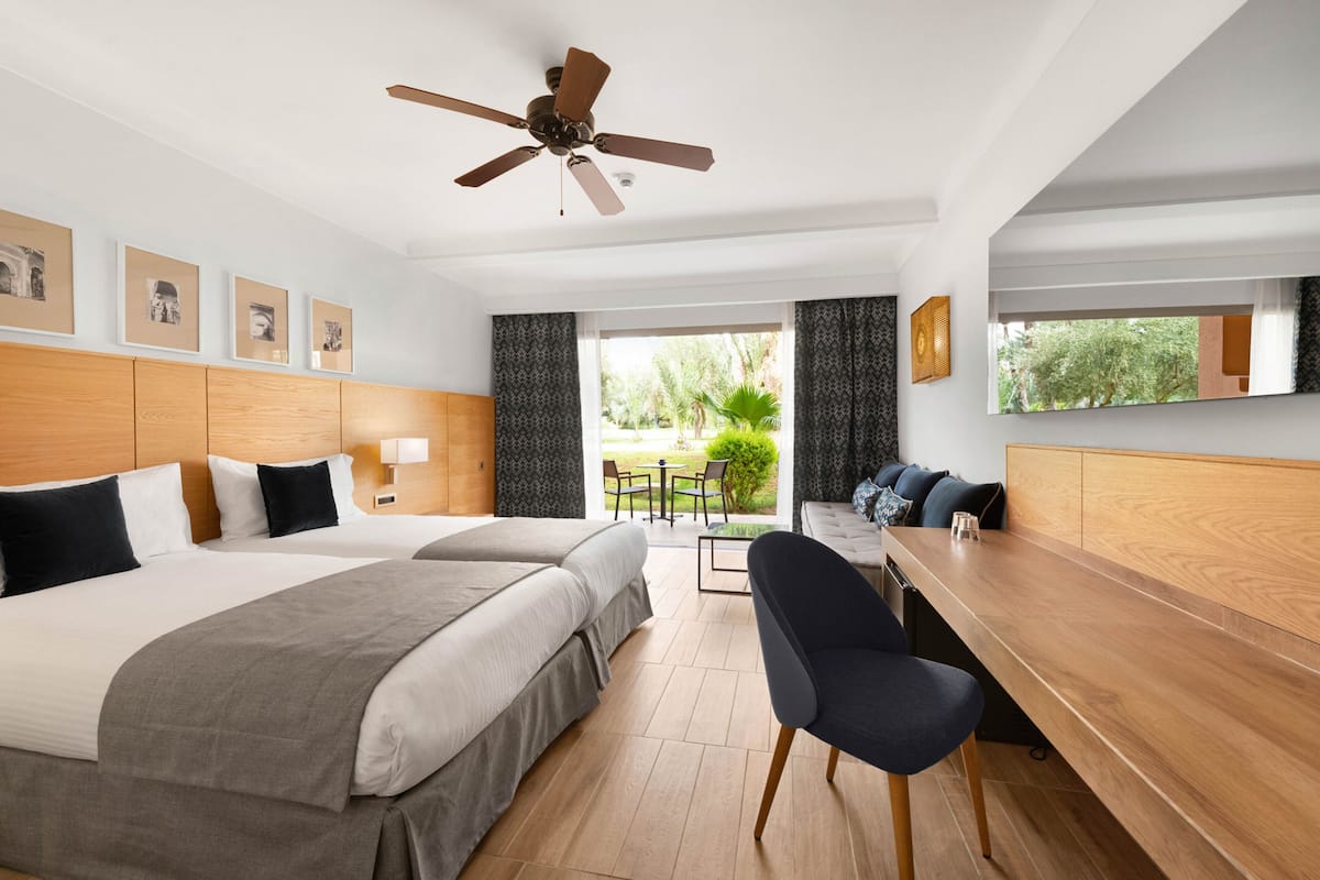 A hotel room with two beds, a desk, a chair, a ceiling fan, and a view of the outdoors.