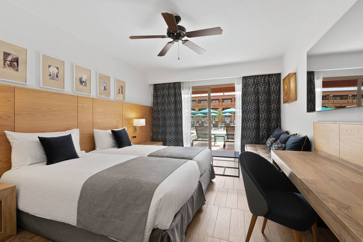 A hotel room with two beds, a desk, a chair, a ceiling fan, and a balcony with outdoor seating.