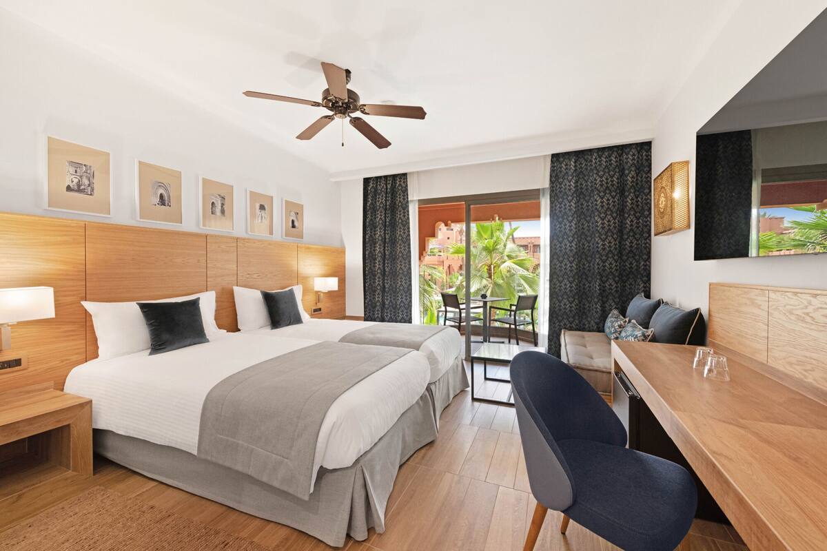 A hotel room with a large bed, a desk, and a balcony with a view of palm trees.