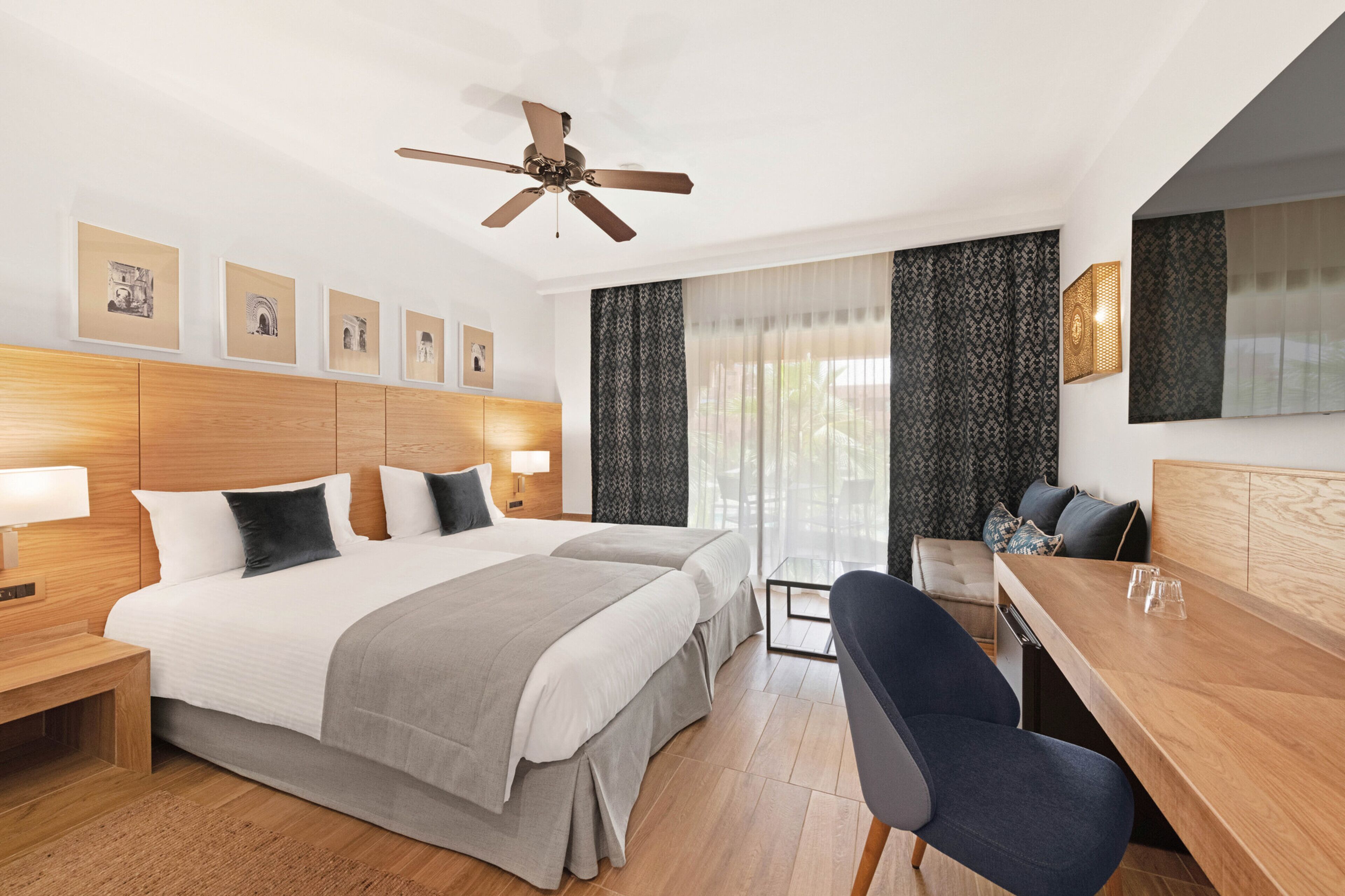 Habitación de hotel con una cama grande, dos mesitas de noche, un escritorio, una silla, un ventilador de techo y una ventana con cortinas.