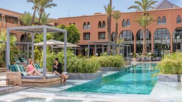 A pool area with lounge chairs and umbrellas, surrounded by palm trees and a large building with arched windows.