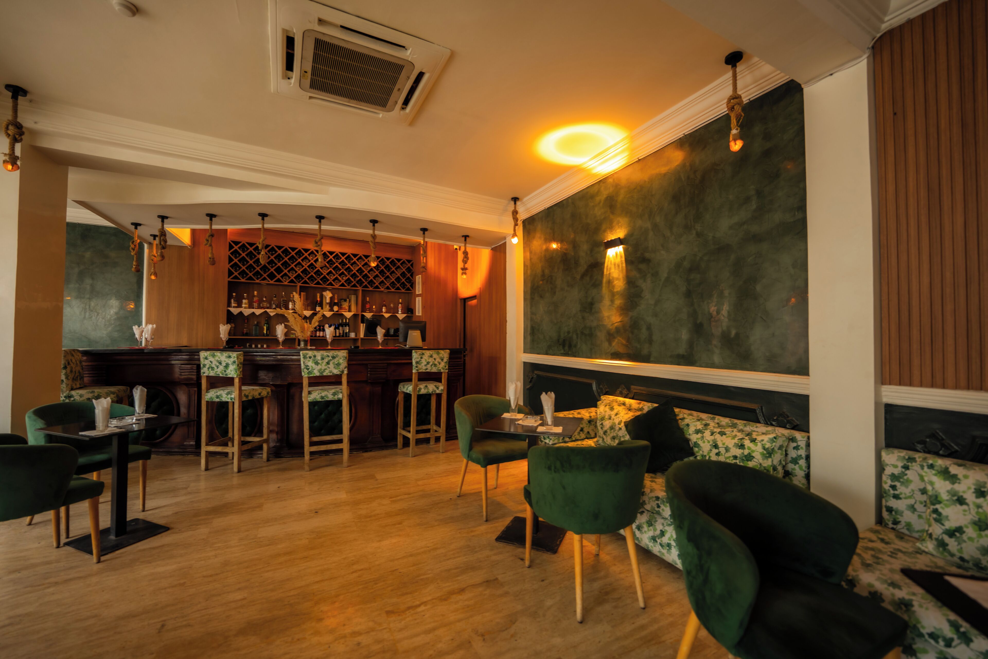 Angolo bar con pavimento in legno, sedute imbottite verdi e bancone bar con bottiglie.
