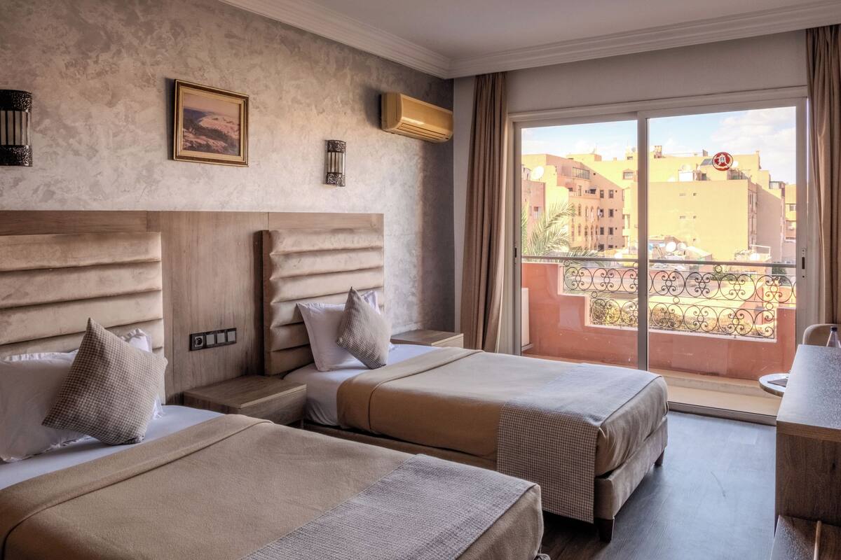 A hotel room with two beds, a large window with a view of buildings, a small table, and a chair.