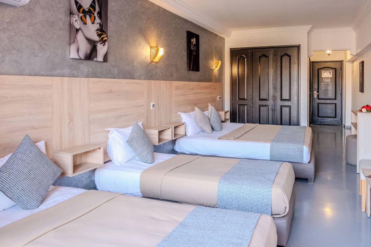 A hotel room with three beds, wooden headboard, wall-mounted light fixtures, and a hallway leading to a door.