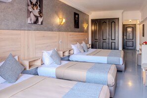 A hotel room with three beds, wooden headboard, wall-mounted light fixtures, and a hallway leading to a door.