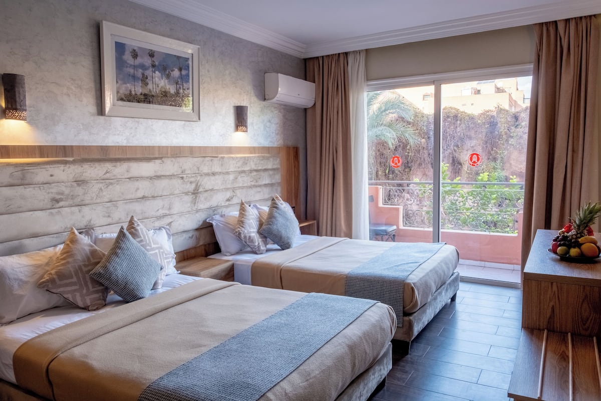A hotel room with two beds, a balcony with a view, and a fruit bowl on a wooden table.