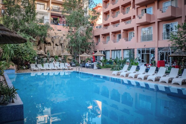 A swimming pool with lounge chairs and umbrellas, surrounded by a building with balconies and vegetation.