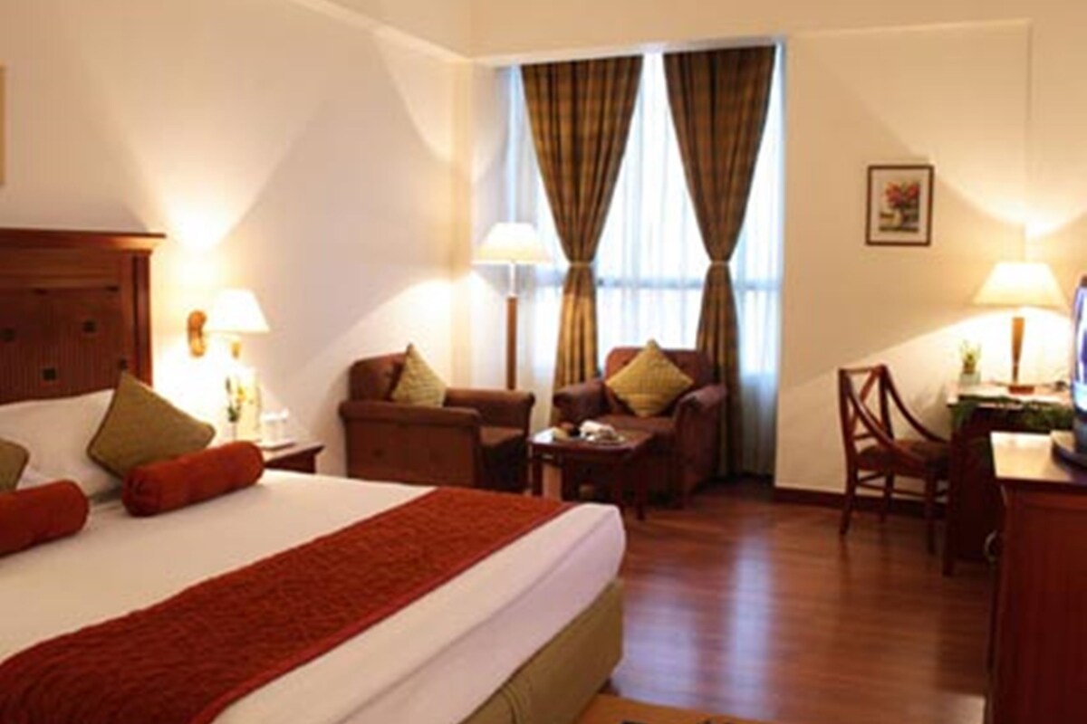 A hotel room with a bed, two armchairs, a desk, and a chair. There are two lamps on the desk and a picture on the wall.