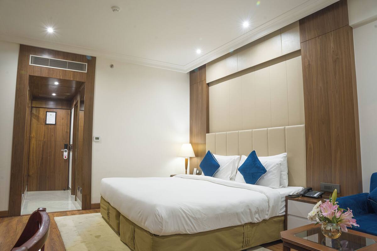 Habitación de hotel con una cama grande, un cabecero de madera, una mesita de noche con una lámpara y una pequeña mesa con un florero.