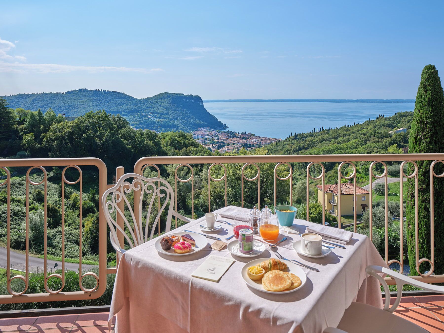 A table set with breakfast items, including pastries, a croissant, and orange juice, overlooking a scenic view of a coastal town and mountains.