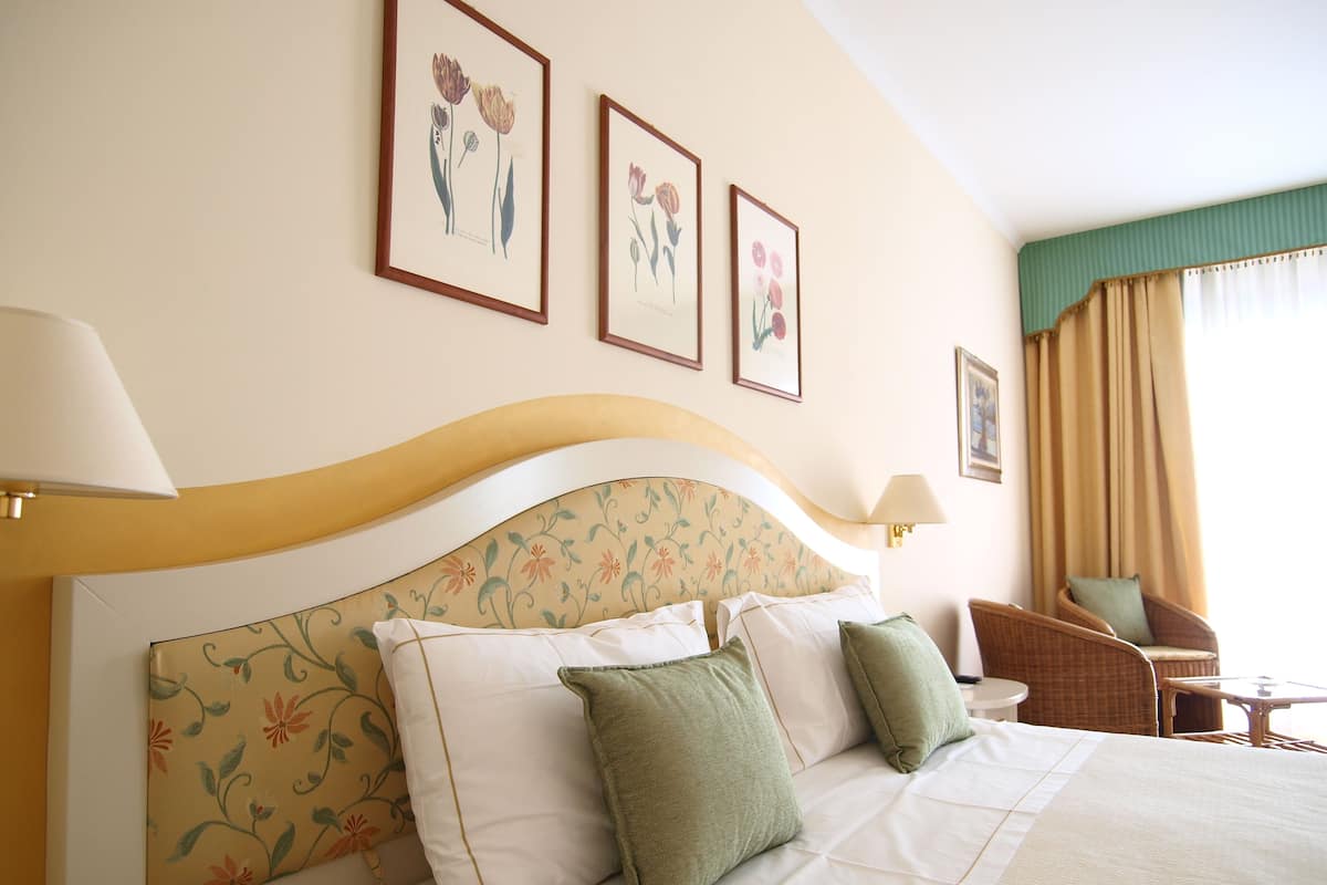 A hotel room with a large bed, floral headboard, green pillows, and framed floral artwork on the wall.