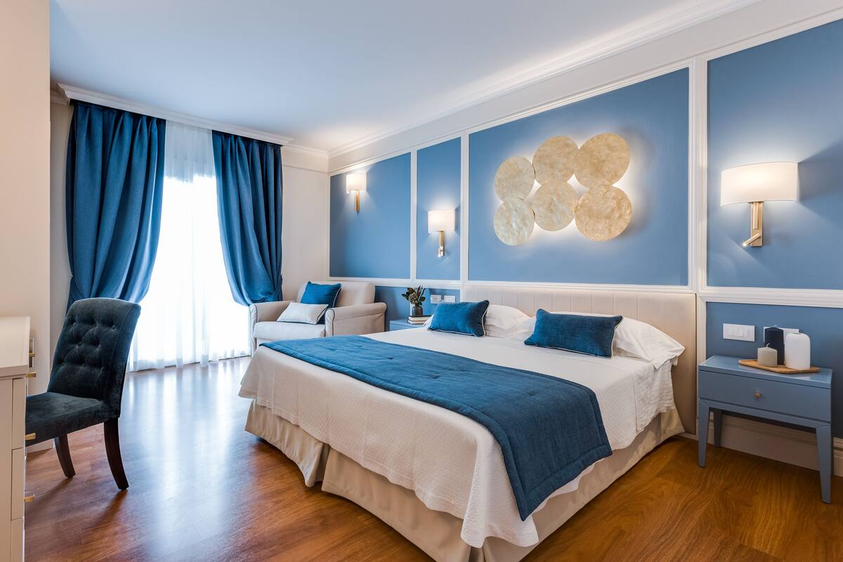 A bedroom with a large bed, blue curtains, a chair, and a nightstand with a lamp.