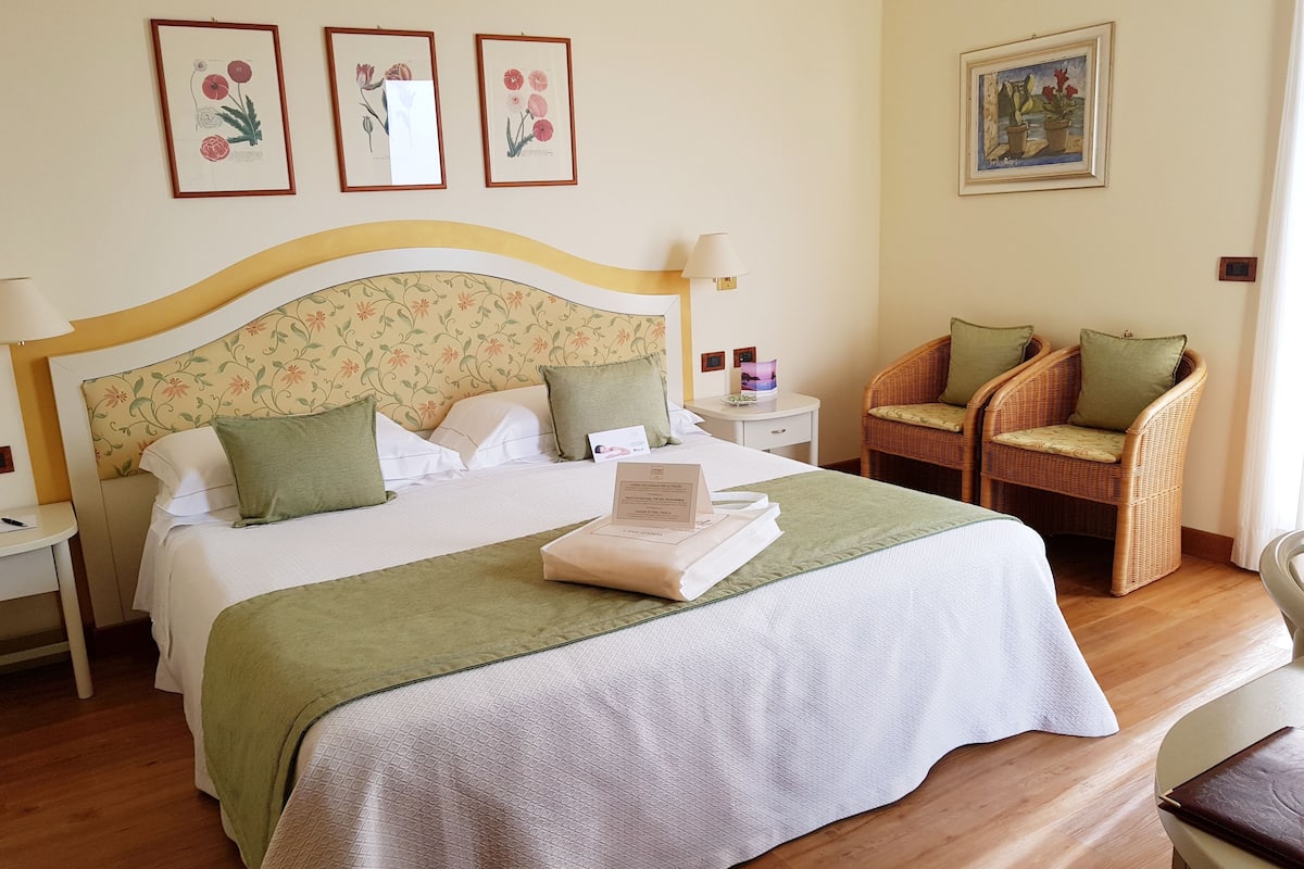 A neatly made bed with a green blanket, a wicker chair with a cushion, and framed botanical prints on the wall.