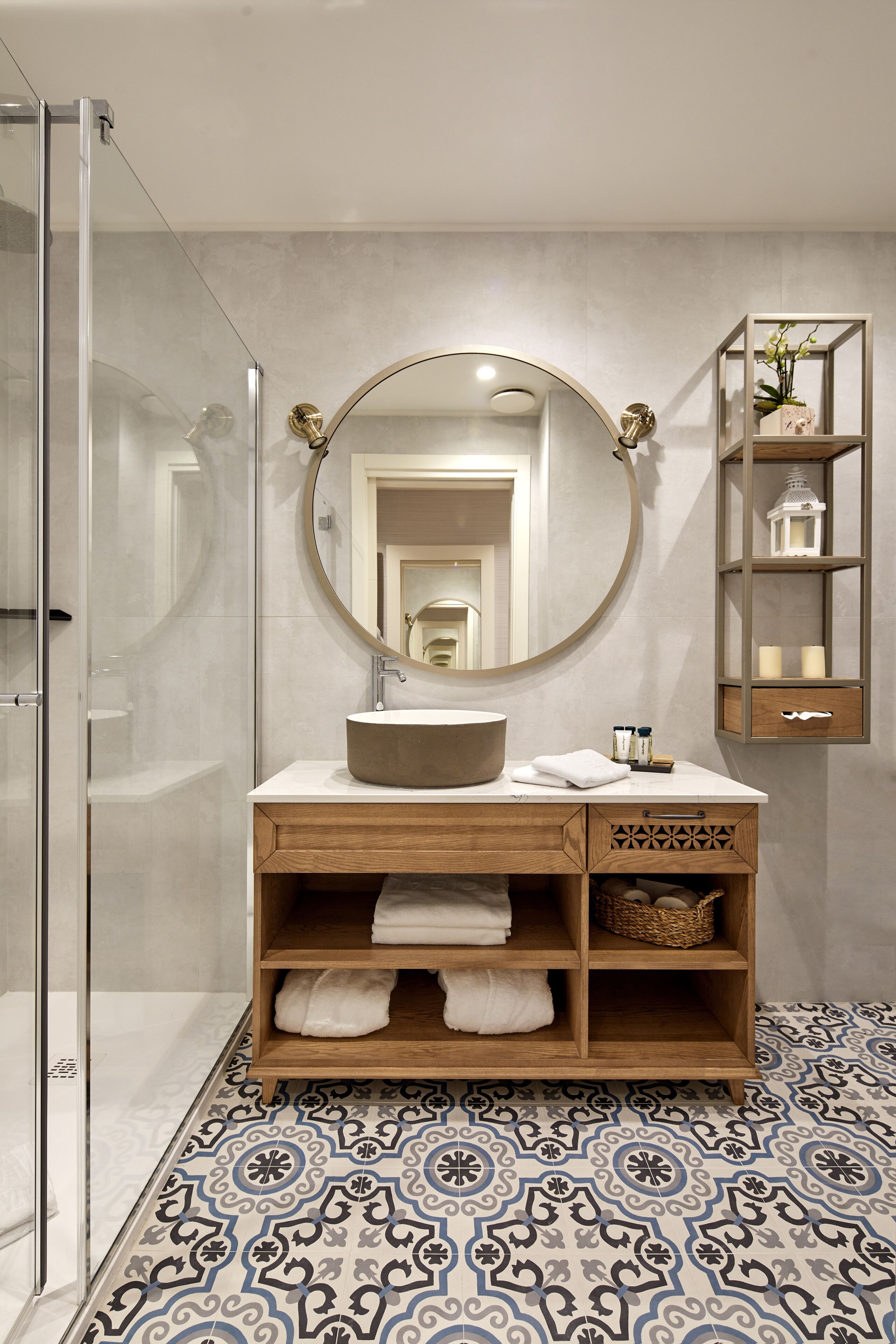 Een moderne badkamer met een glazen douchecabine, een ronde spiegel, een houten wastafelmeubel met een wastafel en vloertegels met een patroon.