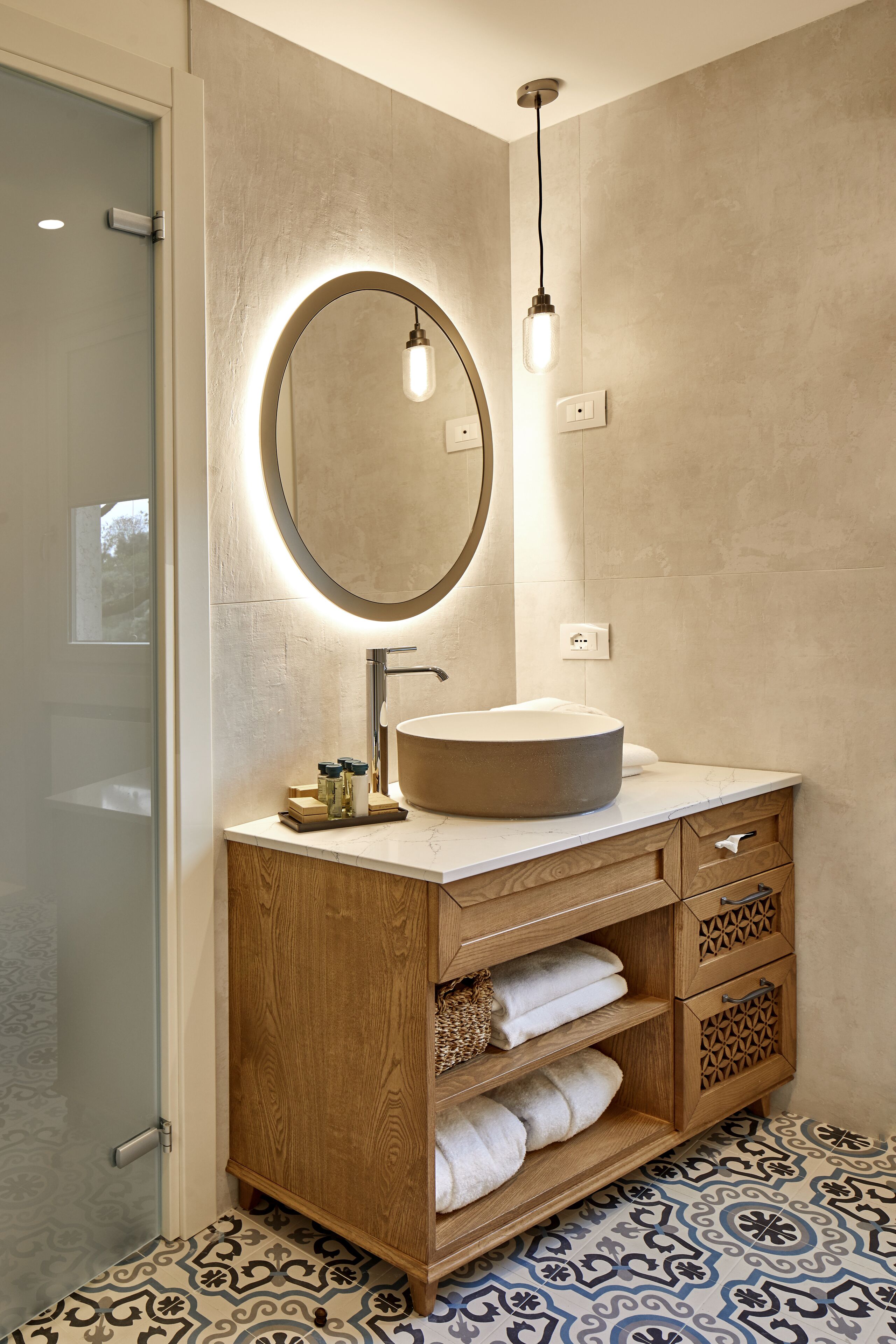 A modern bathroom with a round mirror, a wooden vanity, and patterned floor tiles.