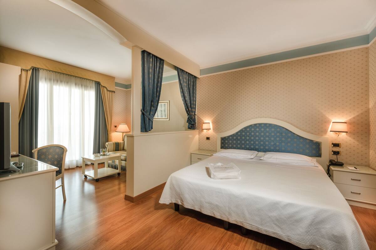 A hotel room with a bed, a desk, a chair, a TV, and a nightstand with a lamp.