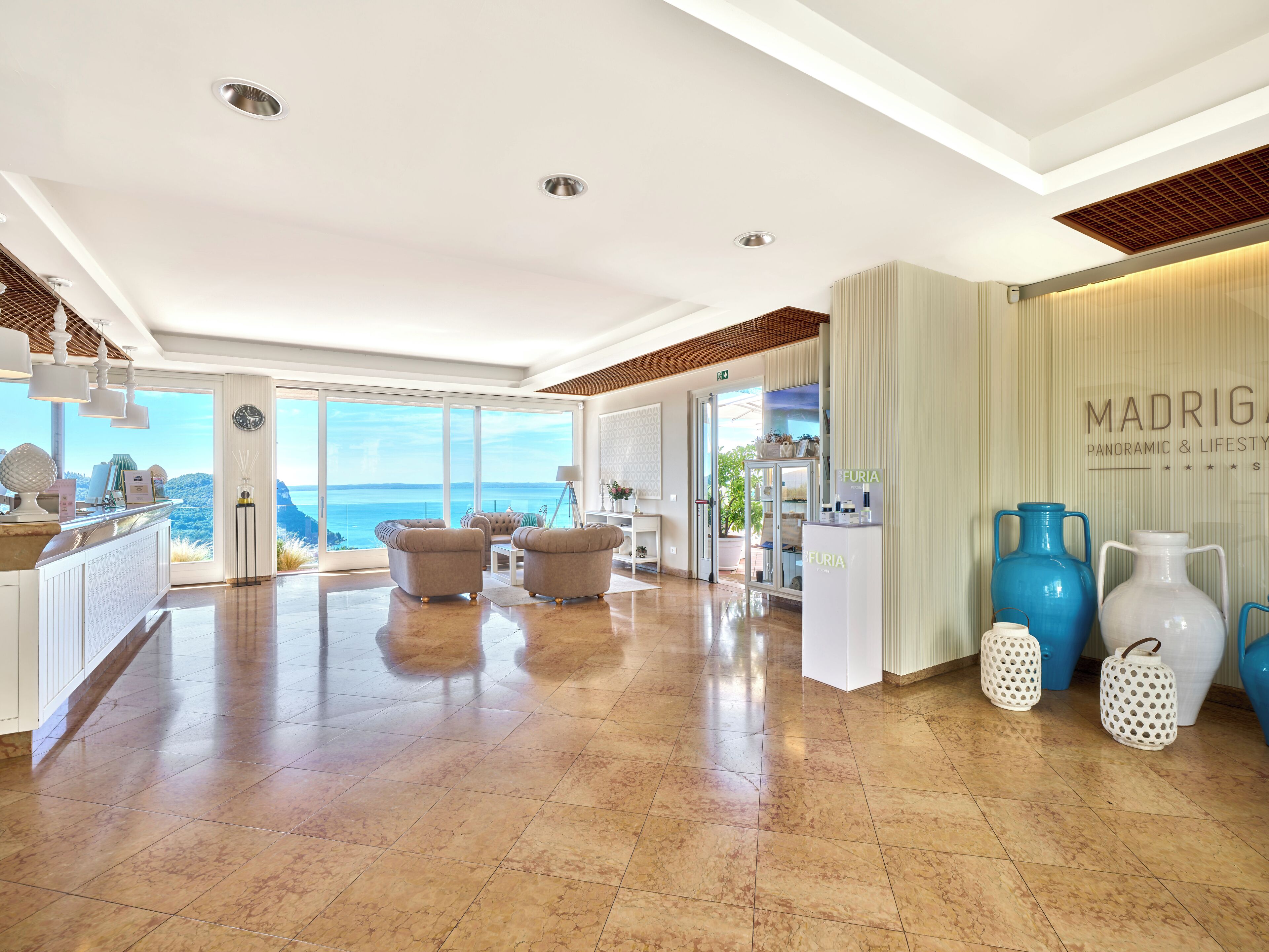 A spacious hotel lobby with a large window offering a view of the sea, a reception desk, and a seating area with two armchairs.