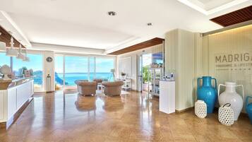 A spacious hotel lobby with a large window offering a view of the sea, a reception desk, and a seating area with two armchairs.
