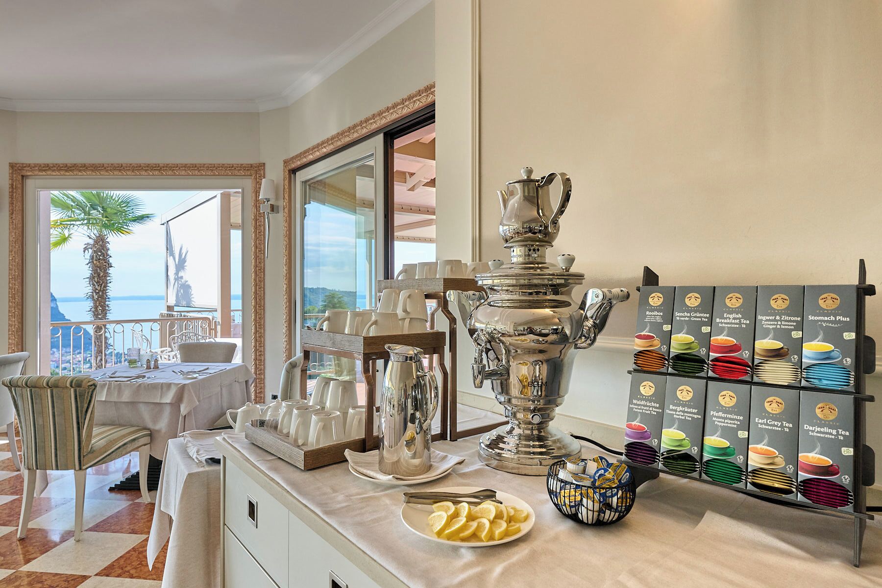 A breakfast buffet setup with a variety of tea options, a large silver teapot, and a view of the sea through a large window.