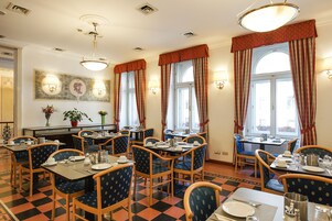 Een restaurantruimte in een hotel met meerdere tafels en stoelen, grote ramen voorzien van gordijnen en wandlampen.