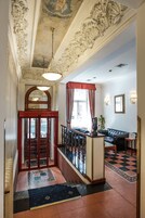 Een statige lobby met decoratieve plafondschilderingen, een vloer met een rood en wit patroon, een zwarte leren bank en een trap met een rode deur.