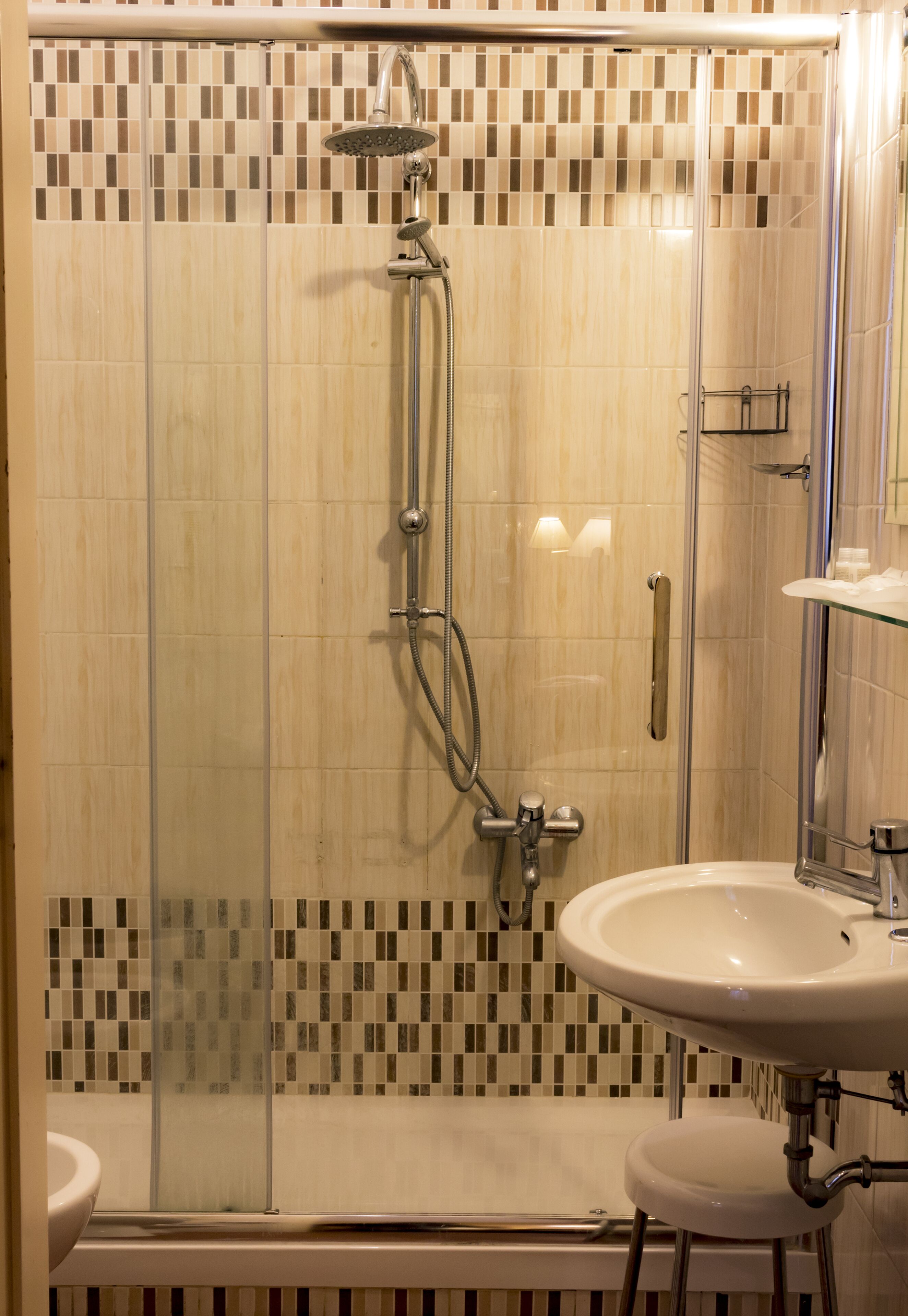 Une douche à l’italienne avec une porte en verre, un lavabo blanc et un mur recouvert de carreaux formant un motif en mosaïque.