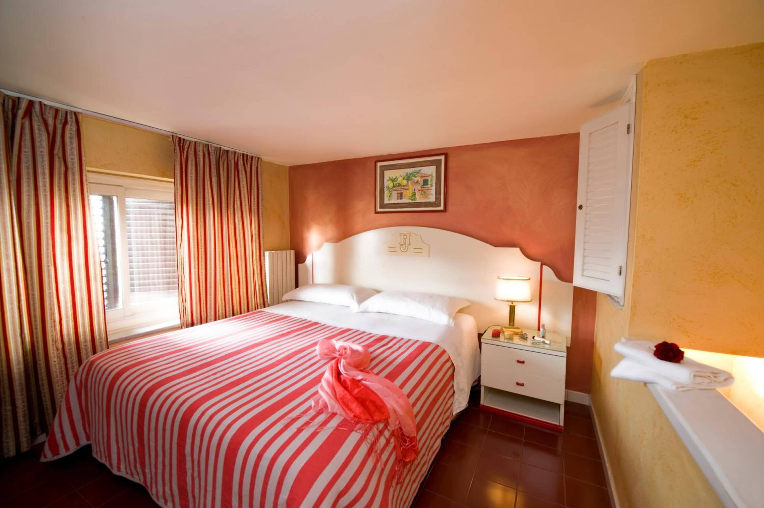 A hotel room with a bed, a nightstand, a lamp, a window with curtains, and a wall with a picture.