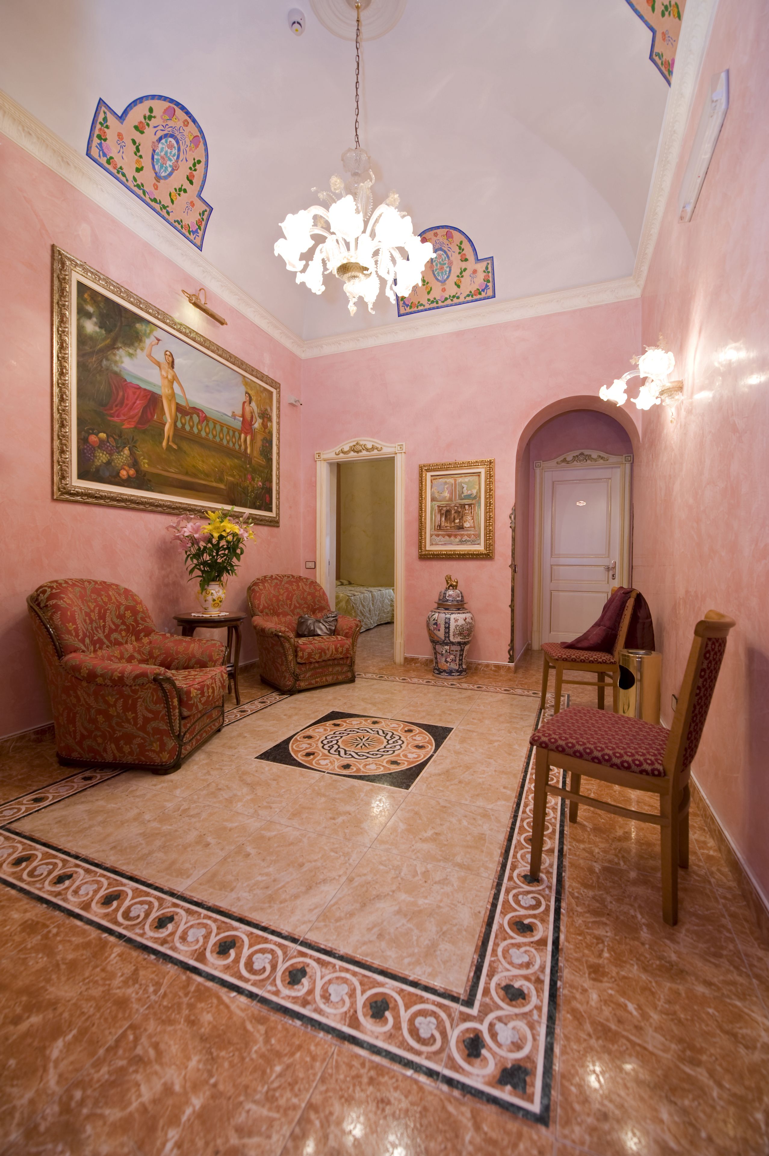 Een kamer met roze muren, een kroonluchter, twee fauteuils, een tafel met een bloevas en een groot schilderij in een lijst.