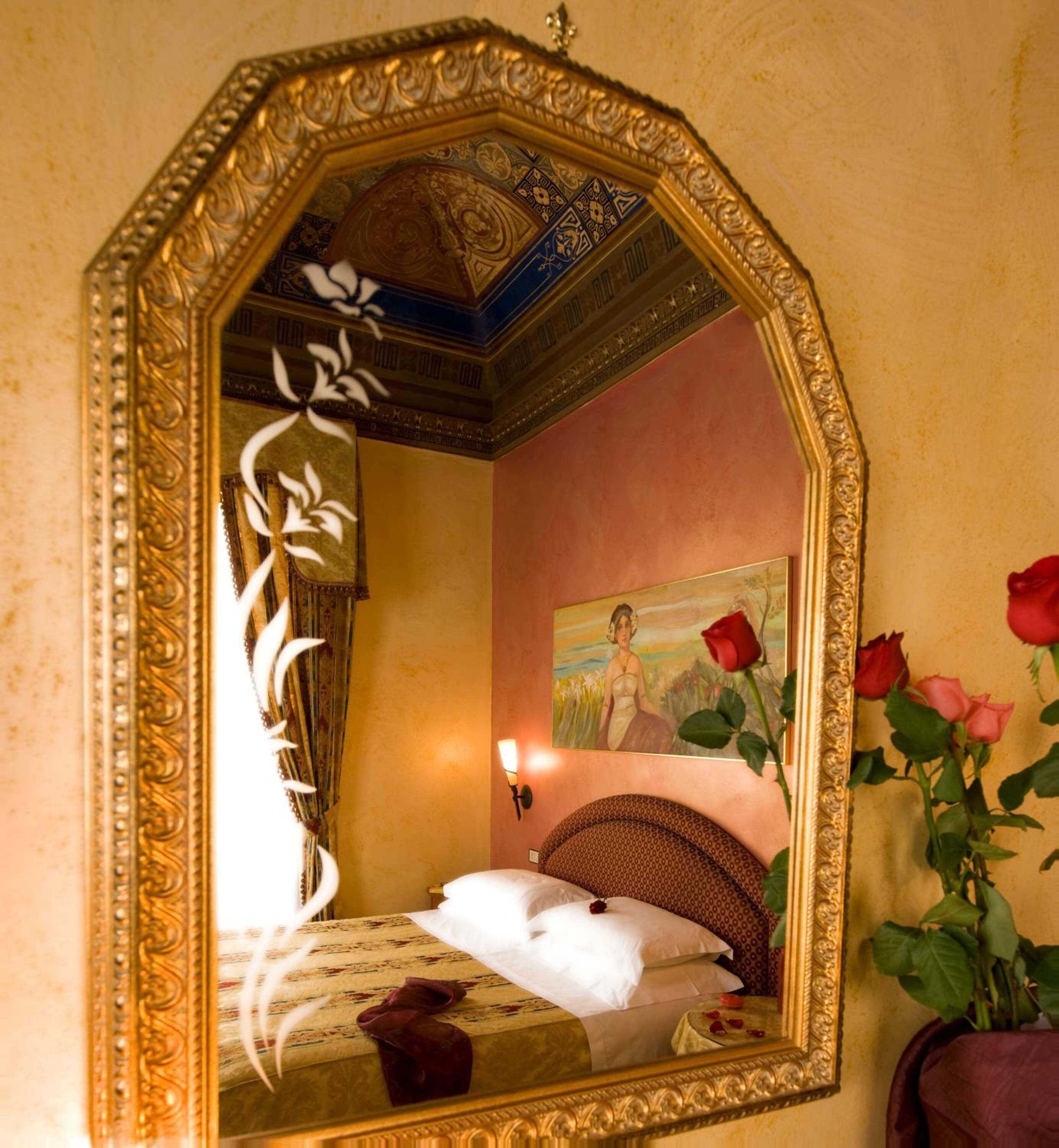 A room with a large ornate mirror, a bed with white linens, a painting on the wall, and a vase with roses.
