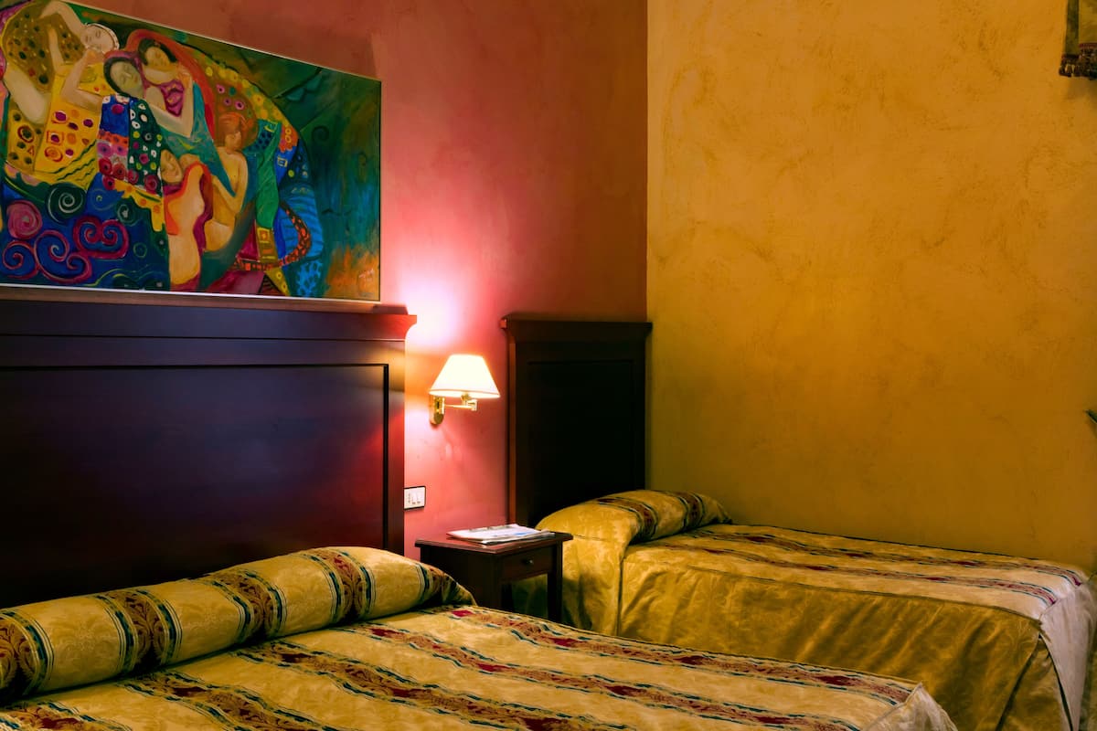Una habitación de hotel con dos camas, un cuadro en la pared, una lámpara y una mesita.