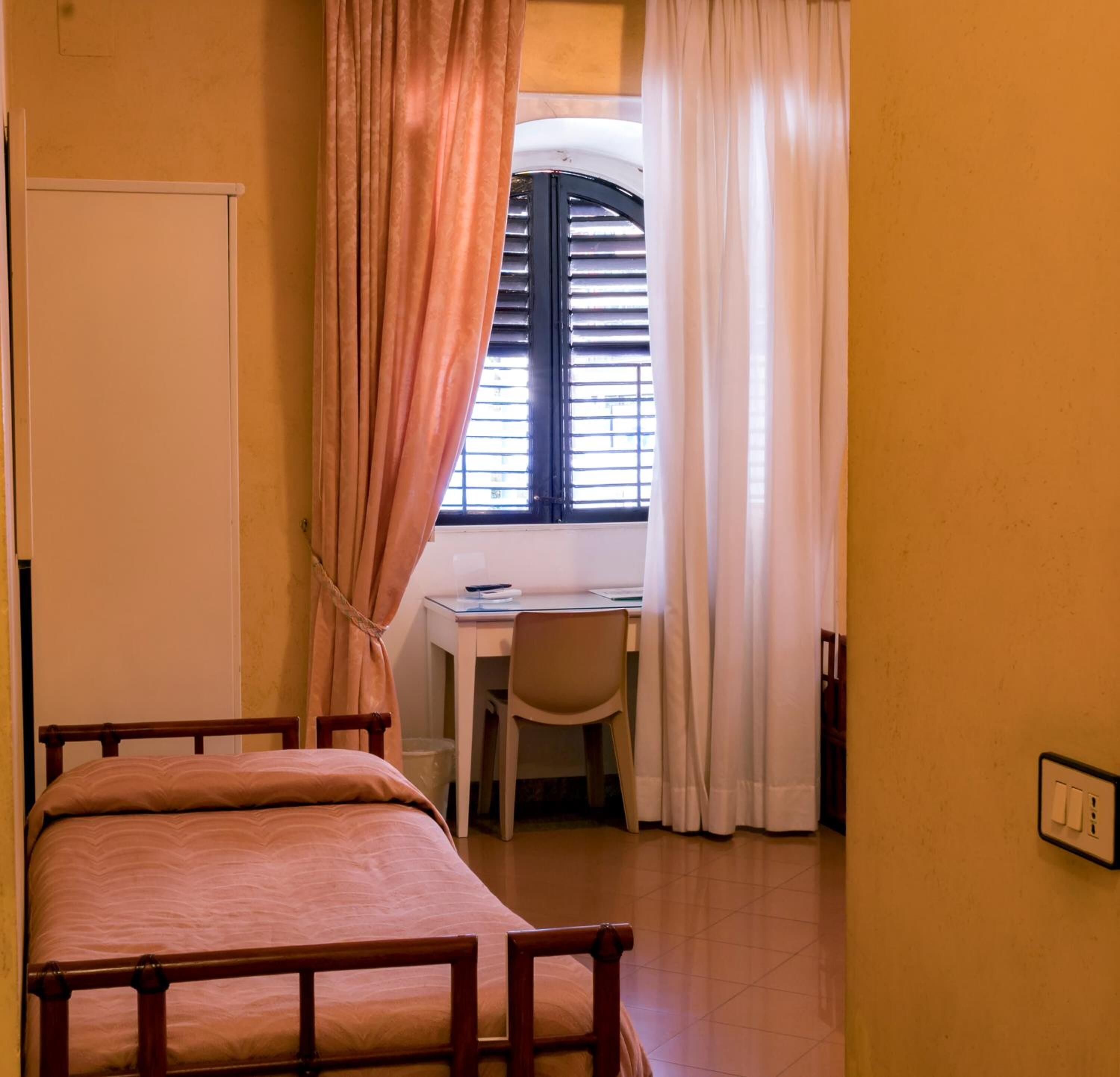 Ein kleines Zimmer mit einem Bett, einem Schreibtisch und einem Stuhl. Ein Fenster mit Jalousien und Vorhängen.
