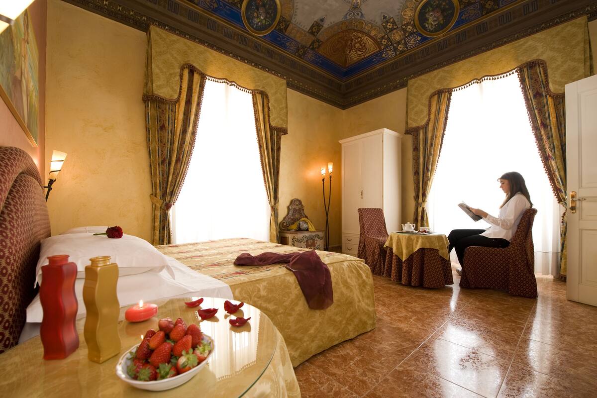 Una habitación de hotel con una cama grande, dos sillones, una mesa con frutero y una mujer leyendo un libro.