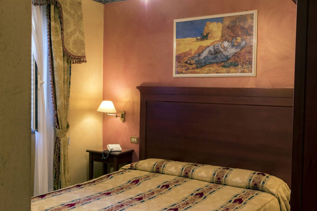 Habitación de hotel con cama, un cuadro en la pared, una lámpara y un teléfono sobre una mesita de noche.