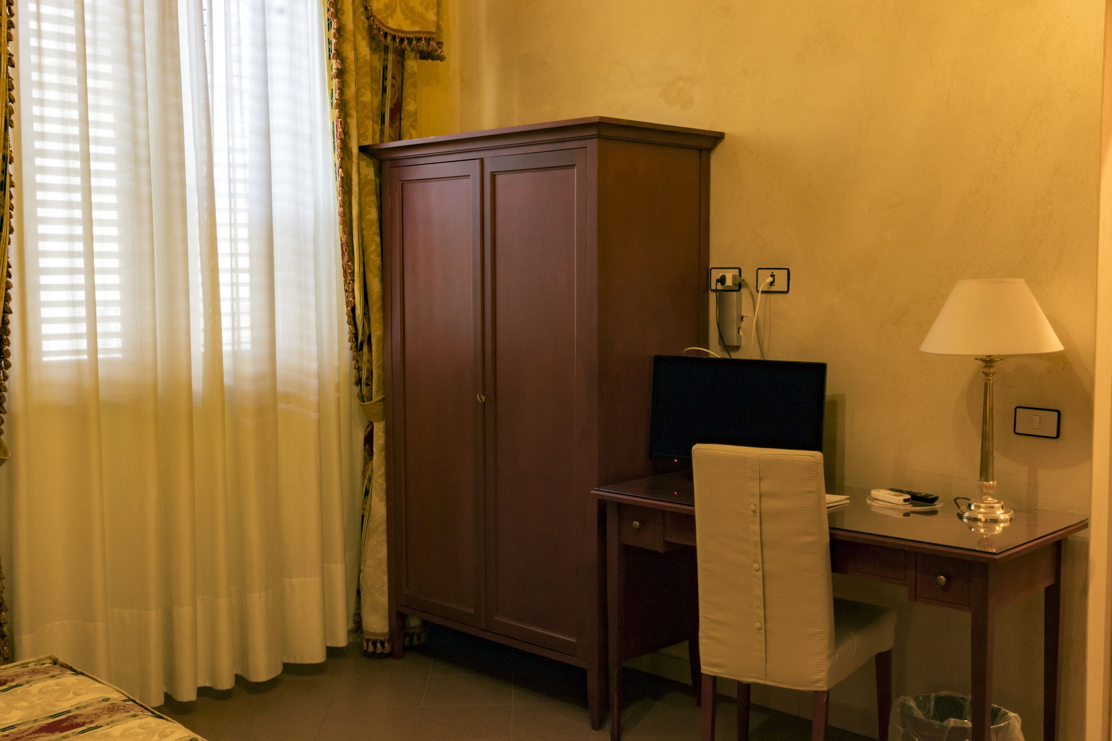 A hotel room with a wooden desk, a chair, a computer, a lamp, and a wardrobe.