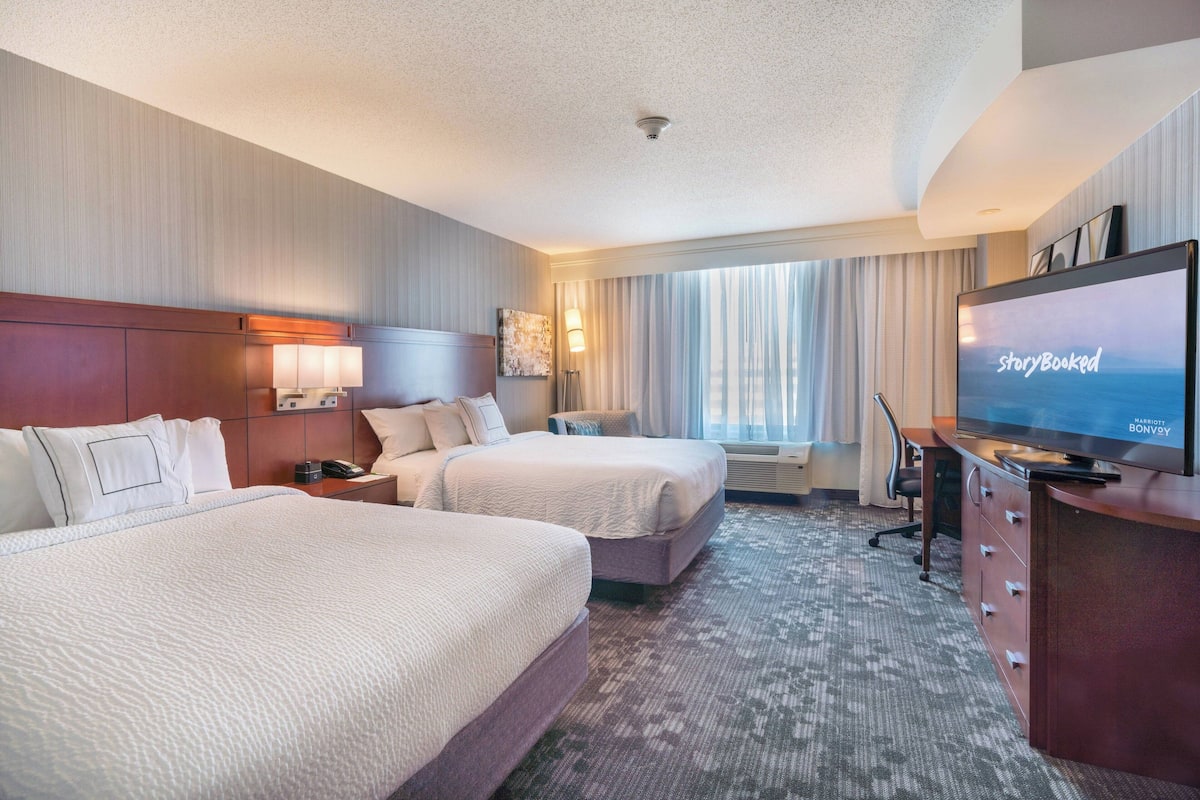 A hotel room with two beds, a television, a desk, and a large window with curtains.
