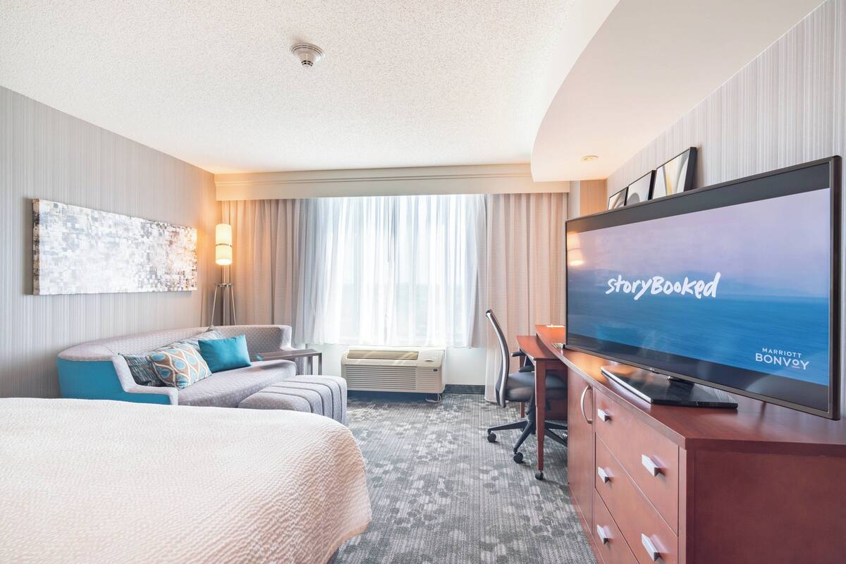 A hotel room with a large bed, a desk, a flat-screen TV, a sofa, and a large window with curtains.