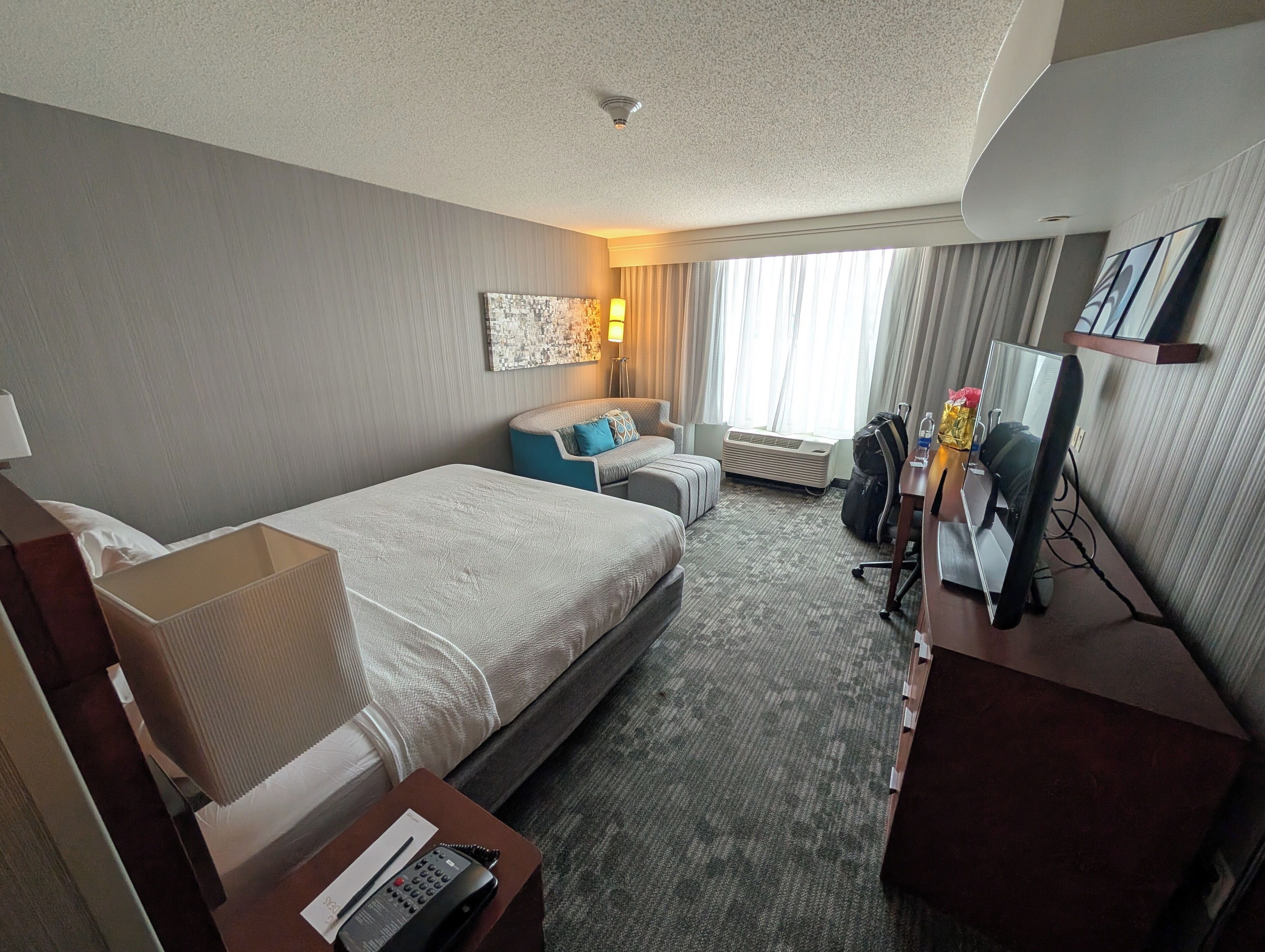 A hotel room with a bed, a desk, a chair, a sofa, a lamp, a painting, and a window with curtains.