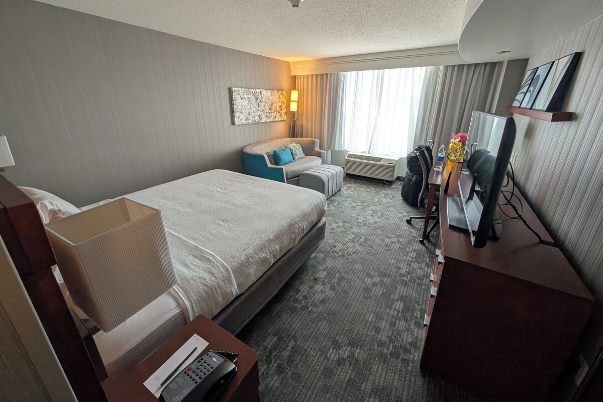 Quarto de hotel com cama, sofá, escrivaninha com computador, luminária e janela com cortinas.