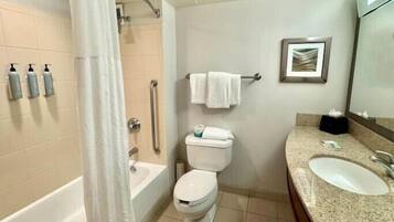 Un baño con bañera, inodoro y lavabo. Hay dispensadores de jabón en la pared, un perchero con toallas y un cuadro enmarcado.