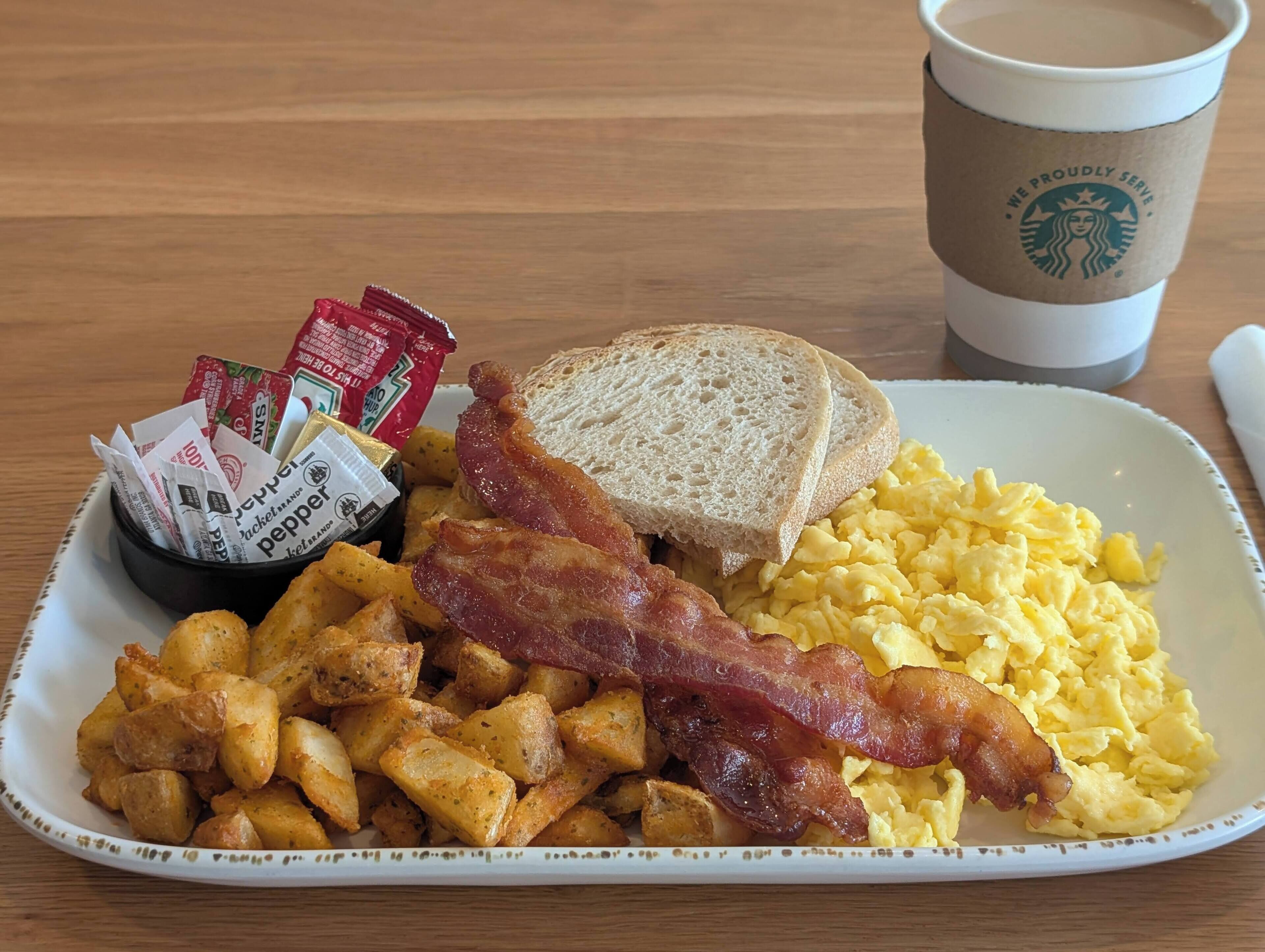 Un plato de desayuno con huevos, tocino, tostadas y papas, junto con una taza de café y salsas.