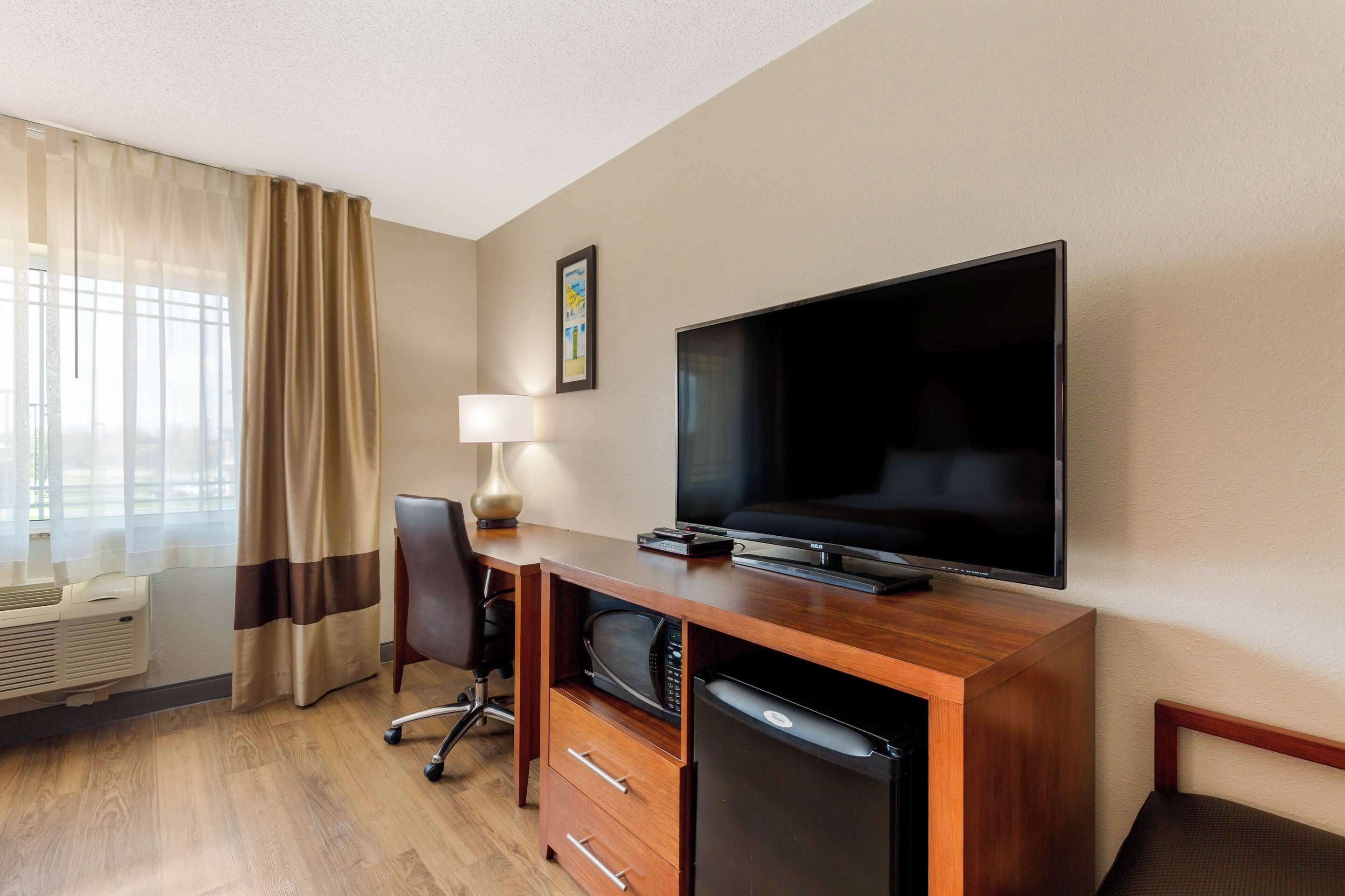 A hotel room with a wooden desk, a flat-screen TV, a lamp, a chair, a mini-fridge, and a window with curtains.