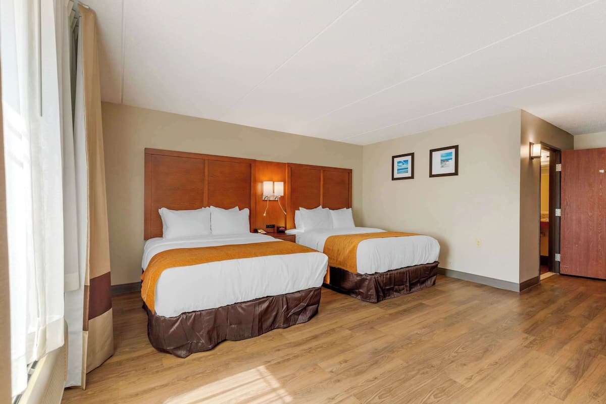 Habitación de hotel con dos camas, cabeceros de madera, una mesita de noche con lámpara y una puerta que da a otra habitación.