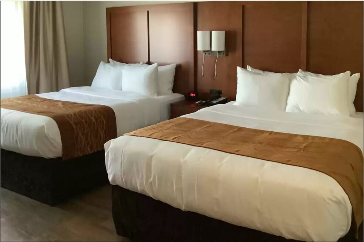 A hotel room with two beds, a nightstand, a wall-mounted lamp, and a clock.