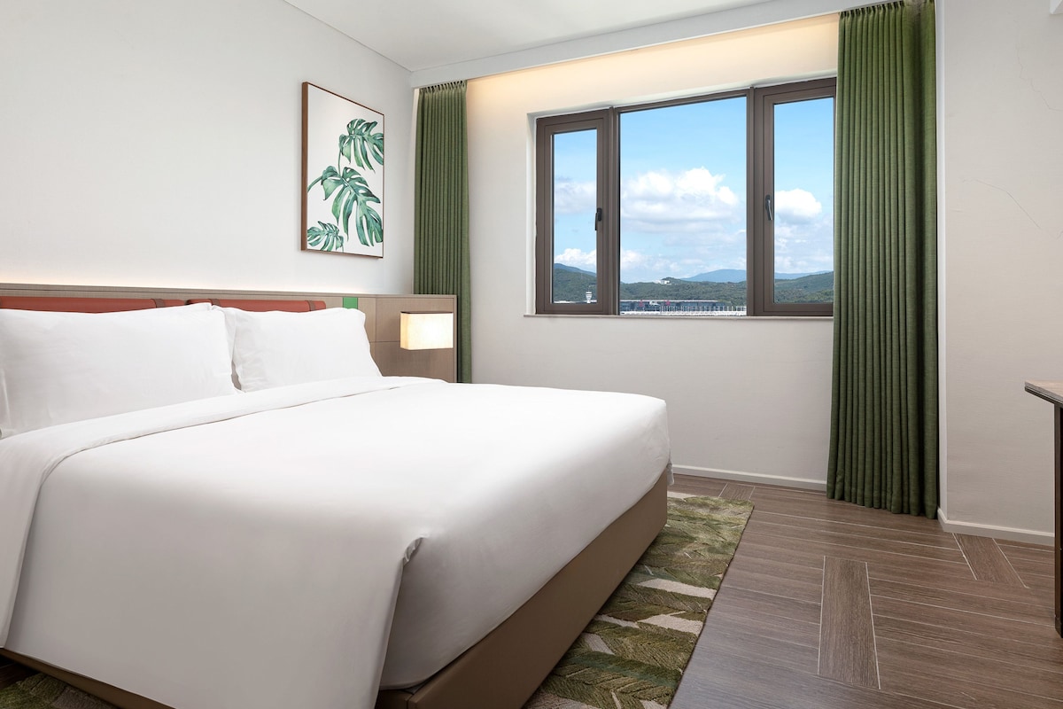A hotel room with a bed, a nightstand, a window with a view, and a painting on the wall.
