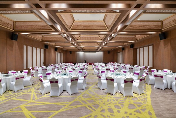 Un salón de banquetes con hileras de mesas redondas, cada una con mantel blanco y fajones morados, y una alfombra estampada amarilla.