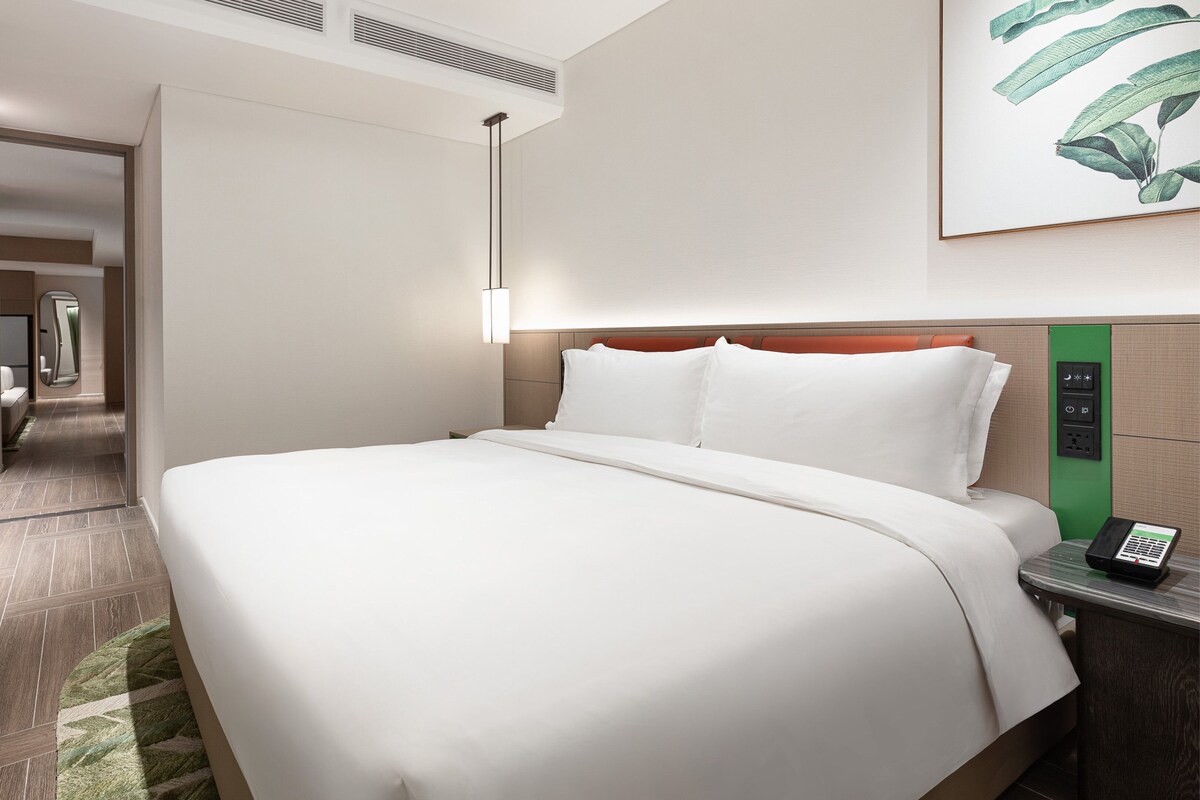 A modern hotel room with a large bed, a bedside table with a phone, a wall-mounted control panel, and a hallway leading to a bathroom.