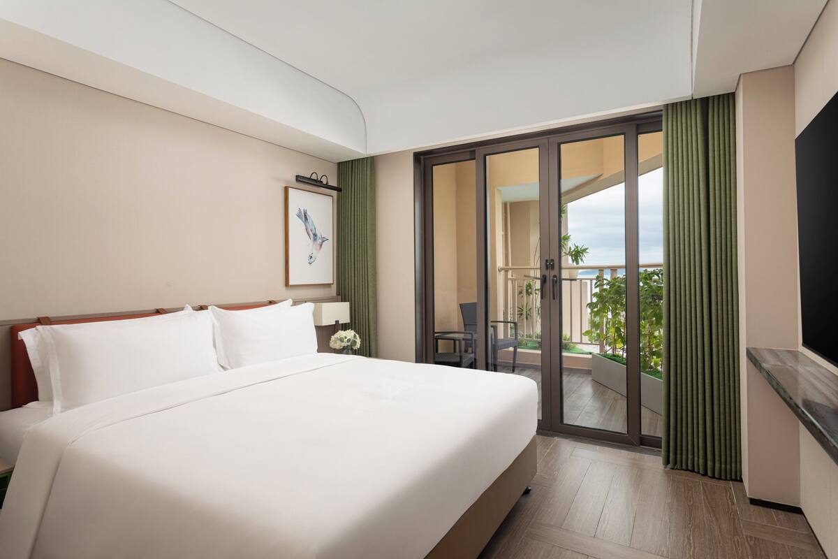 Ein Hotelzimmer mit einem großen Bett, einem Fernseher, einer Schiebetür zu einem Balkon und einem gerahmten Bild an der Wand.