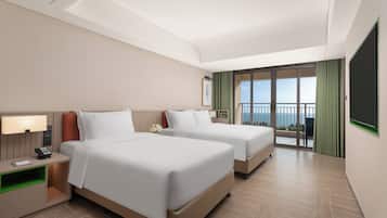 Quarto de hotel com duas camas, uma mesinha de cabeceira, escrivaninha, uma cadeira e uma janela grande com vista para o oceano.